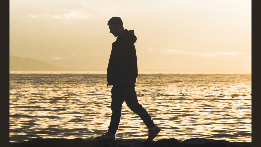 Homem caminha reflexivo na beira do mar.