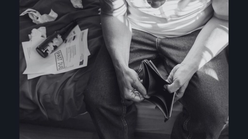 Homem sentado na cama, olhando uma carteira vazia, enquanto contas atrasadas e papéis financeiros estão espalhados ao lado — retratando a pressão e a responsabilidade sobre a vida financeira masculina.