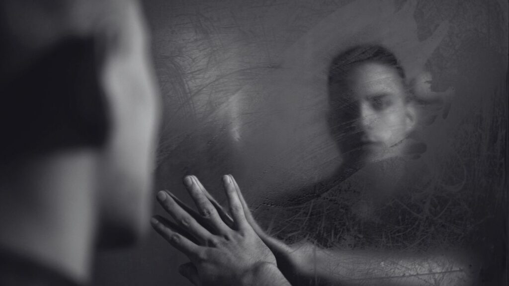 Homem olhando para o próprio reflexo em um espelho embaçado, com a mão apoiada no vidro — simbolizando conflito interno, consciência e reflexão sobre a própria vida e decisões.