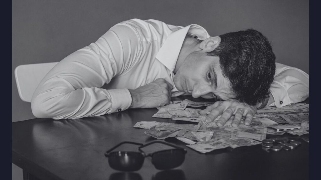 Homem deitado sobre a mesa, com expressão cansada, cercado por notas de dinheiro e moedas espalhadas ,simbolizando desgaste emocional e descontrole na vida financeira.